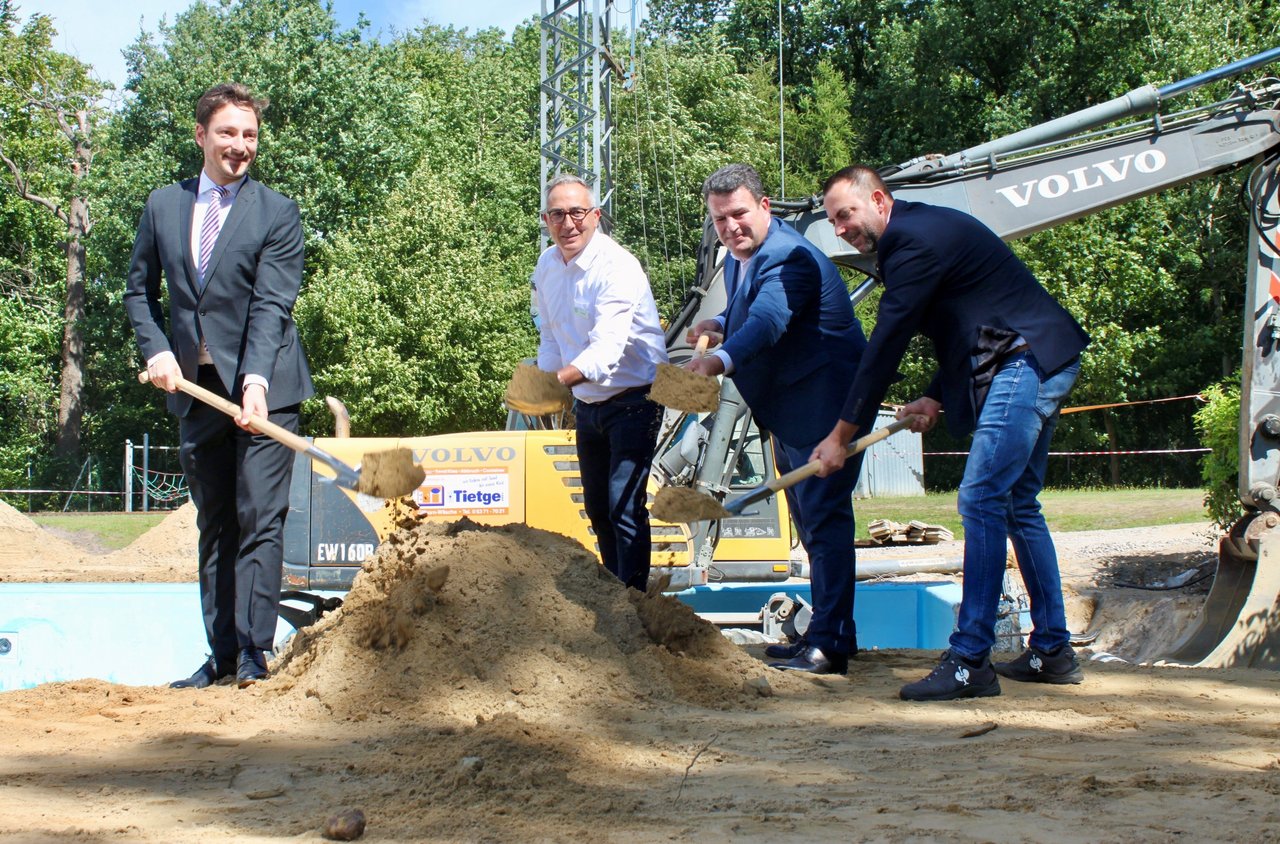 Man sieht vier Männer im Anzug, die einen Spaten mit Sand in die Luft heben.