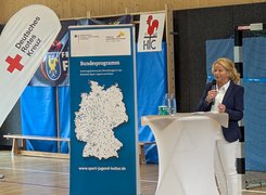 Quelle: Stadt Goslar Das Bild zeigt eine Frau mit Mikrofon an einem Stehtisch, im Hintergrund sind mehrere Aufsteller und Sportgeräte zu sehen.