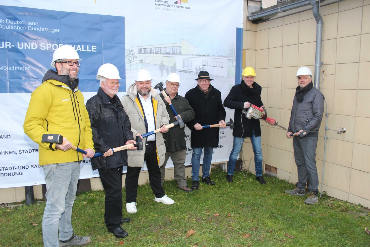 Man sieht sieben Männer, die einen Bauhelm tragen und einen Hammer in der Hand halten, vor einem Bauschildplakat.