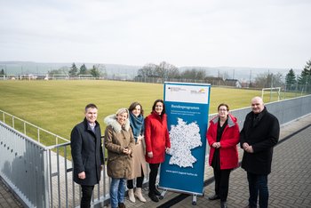 Man sieht sechs Personen, die an einem Fußballplatz neben einem Roll-Up stehen.