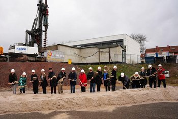 Das Bild zeigt eine Reihe von Personen mit weißem Bauhelm, die alle einen Spaten in der Hand halten. Dies symbolisiert den Baubeginn an einer Baustelle. Im Hintergrund ist ein Bauzaun und Bagger zu sehen.