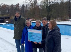 Man sieht fünf Personen in einem verschneiten Freibad. die einen großen Spendenscheck halten. 