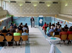 Schwimmbads. Quelle: Gemeinde Türkenfeld Das Bild zeigt eine Person am Rednerpult vor Menschen in Stuhlreihen sitzend, das Setting ist ein leeres Schwimmbecken.