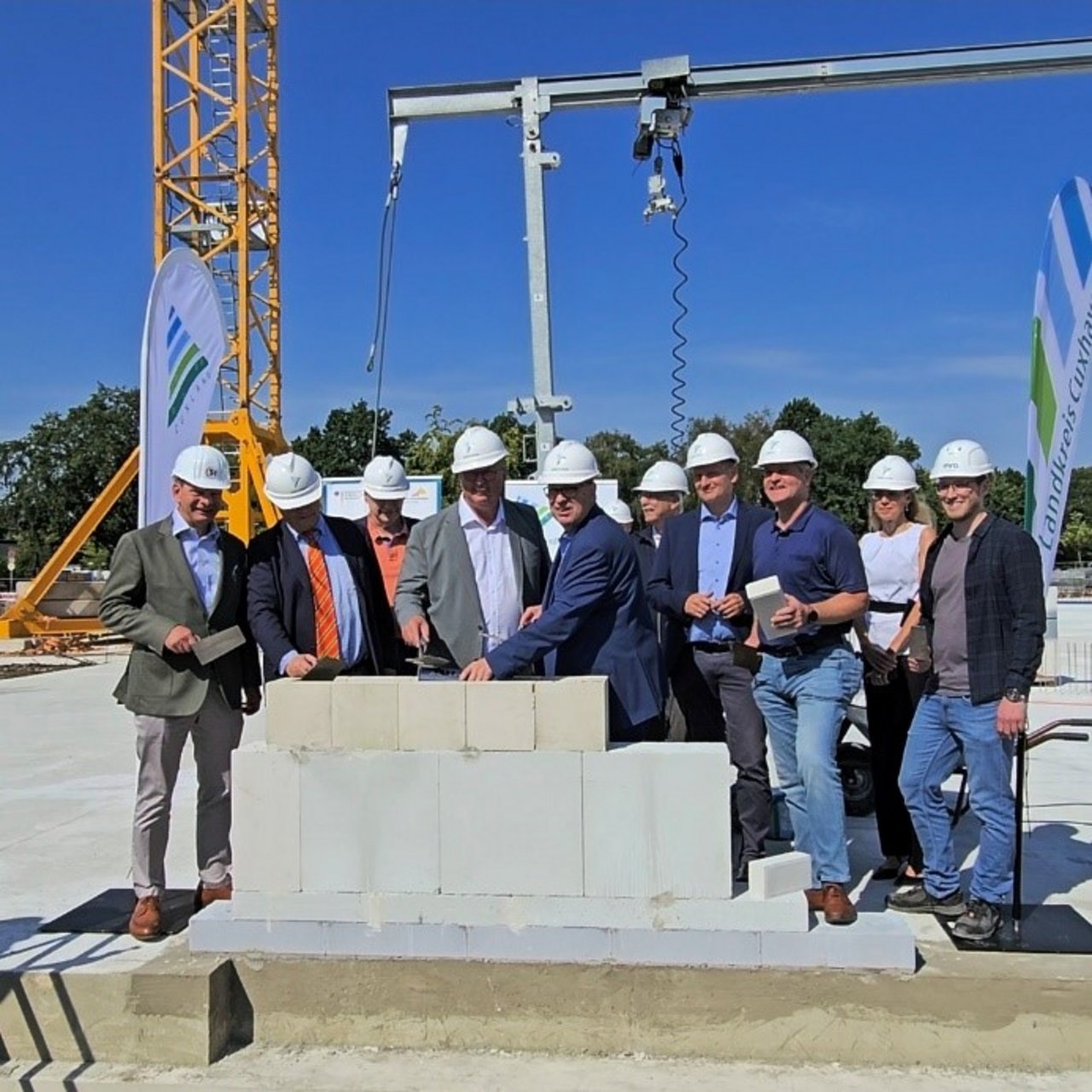 Man sieht viele Menschen, die Bauhelme tragen und vor einem Kran an einer teilweise gebauten Mauer stehen.