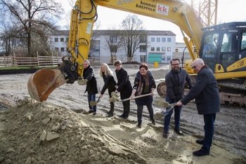 Auf dem Foto sind sechs Personen zu sehen. Sie stehen vor einem Bagger und halten eine Schaufel in der Hand. 