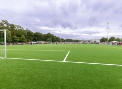 Das Bild zeigt einen Fußballplatz, wo ein Spiel ausgetragen wird. Zuschauer stehen um das Spielfeld herum.