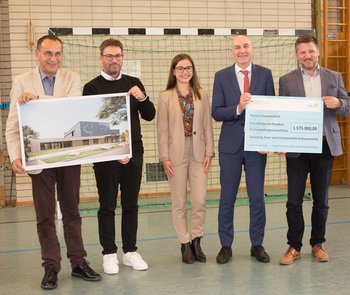 Man sieht vier Männer und eine Frau, die zwei Spendenschecks hoch halten und vor einem Tor in einer Sporthalle stehen.