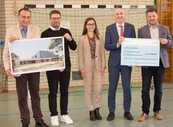 Man sieht vier Männer und eine Frau, die zwei Spendenschecks hoch halten und vor einem Tor in einer Sporthalle stehen.