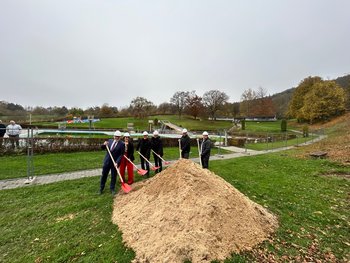 Man sieht einen großen Sandhaufen hinter dem sechse Perosnen mit Spaten und Bauhelmen stehen.