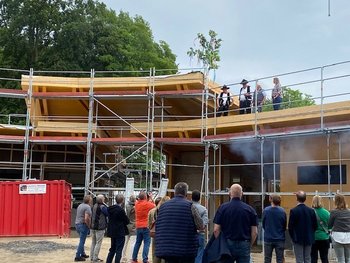 Das Bild zeigt eine Baustelle mit einem Rohbau und einem Gerüst. Auf und vor dem Gerüst befinden sich Personen bei einem Richtfest.