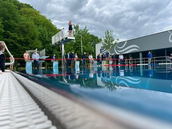 Das Bild zeigt mehrere Personen, die vor dem Schwimmbecken stehen. Eine Person befindet sich mit verschränktem Armen auf einem Sprungbrett.