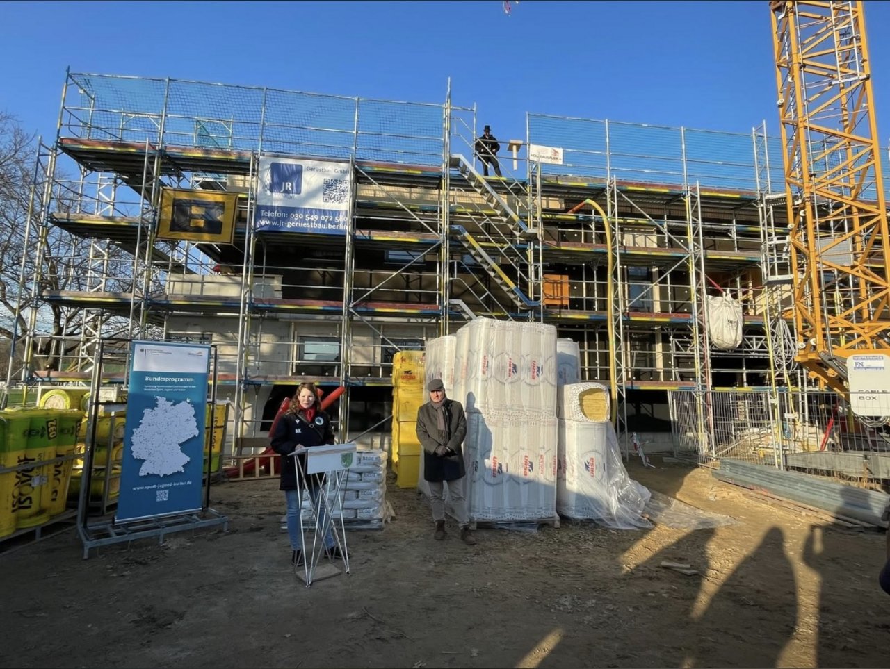 Man sieht zwei Menschen vor einem eingerüsteten Gebäude auf einer Baustelle. Sie halten eine Rede. 