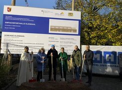 Quelle: Gemeinde Kleinkahl Man sieht sechs Personen mit Spaten die vor einem Bauschild zur Einfachsporthalle Kleinkahl stehen.