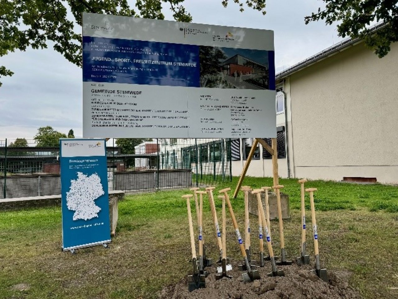 Man sieht viele Spaten, die in einem Erdhaufen vor einem Bauschild und einem Roll-Up stehen.