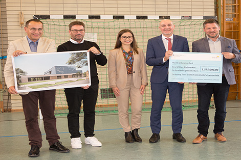 Das Bild zeigt 5 Personen, zwei halten ein Konzeptbild der neuen Turn- und Schwimmhalle und zwei andere halten einen Fördercheck 